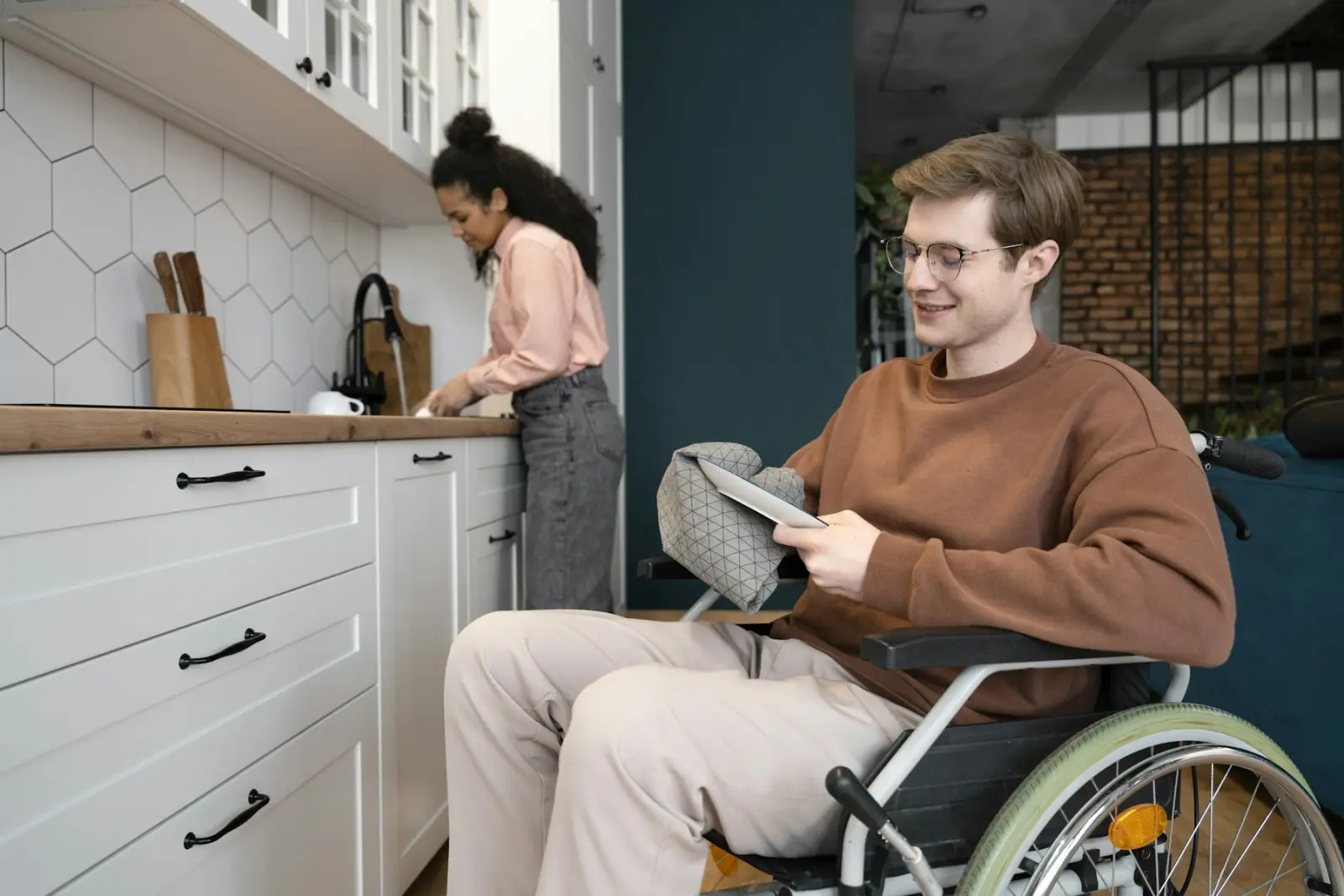 Een jongeman in een rolstoel in zijn eigen keuken, met een begeleider op de achtergrond